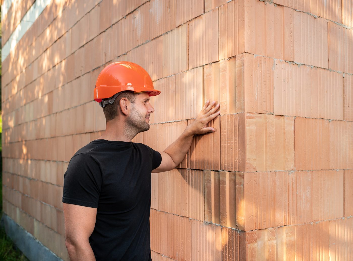 Muž stojící před cihlovou stěnou z cihel Porotherm T Profi na hlavě s oranžovou helmou rukou hladící cihly