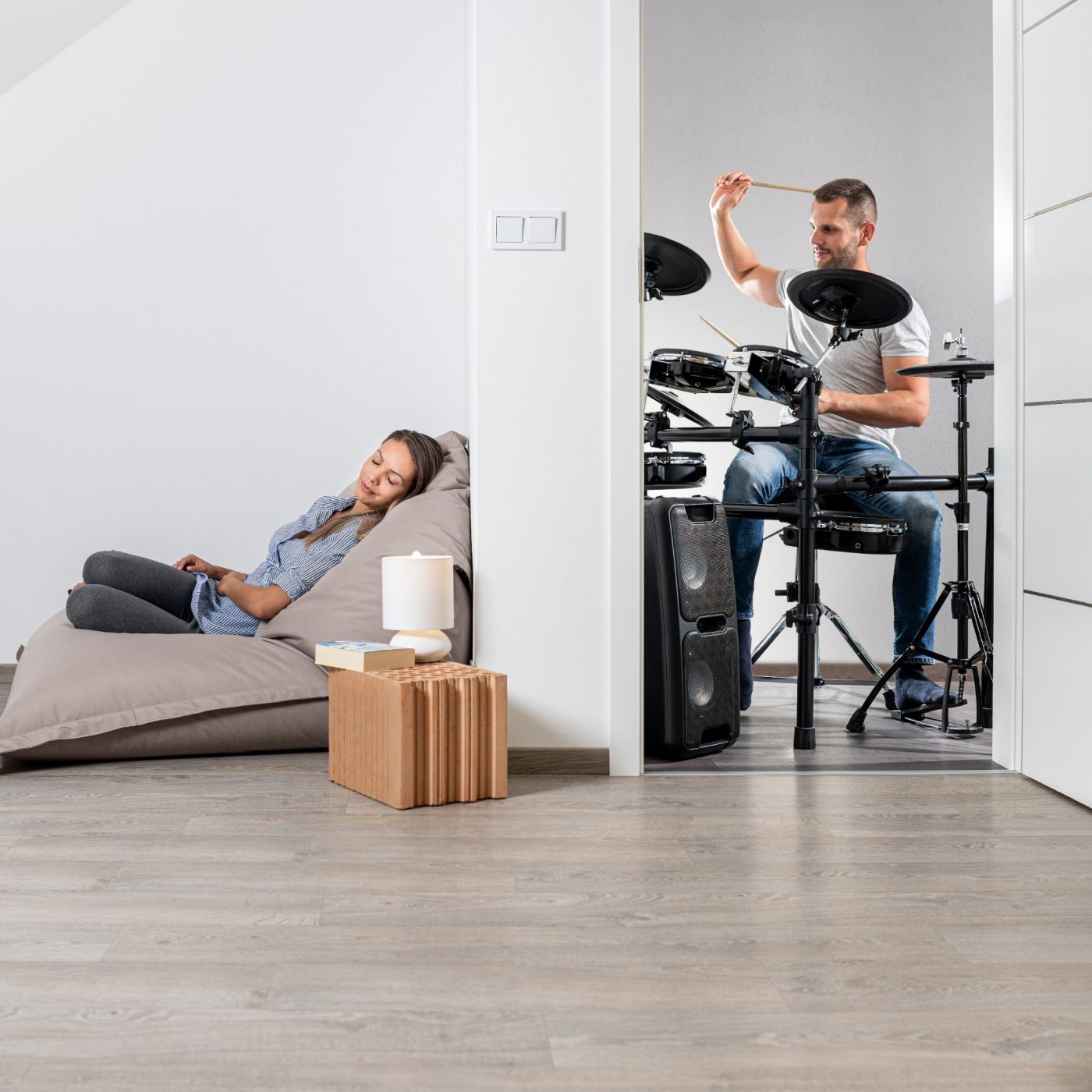 Muž hrající na bicí a v druhé místnosti klidně ležící žena