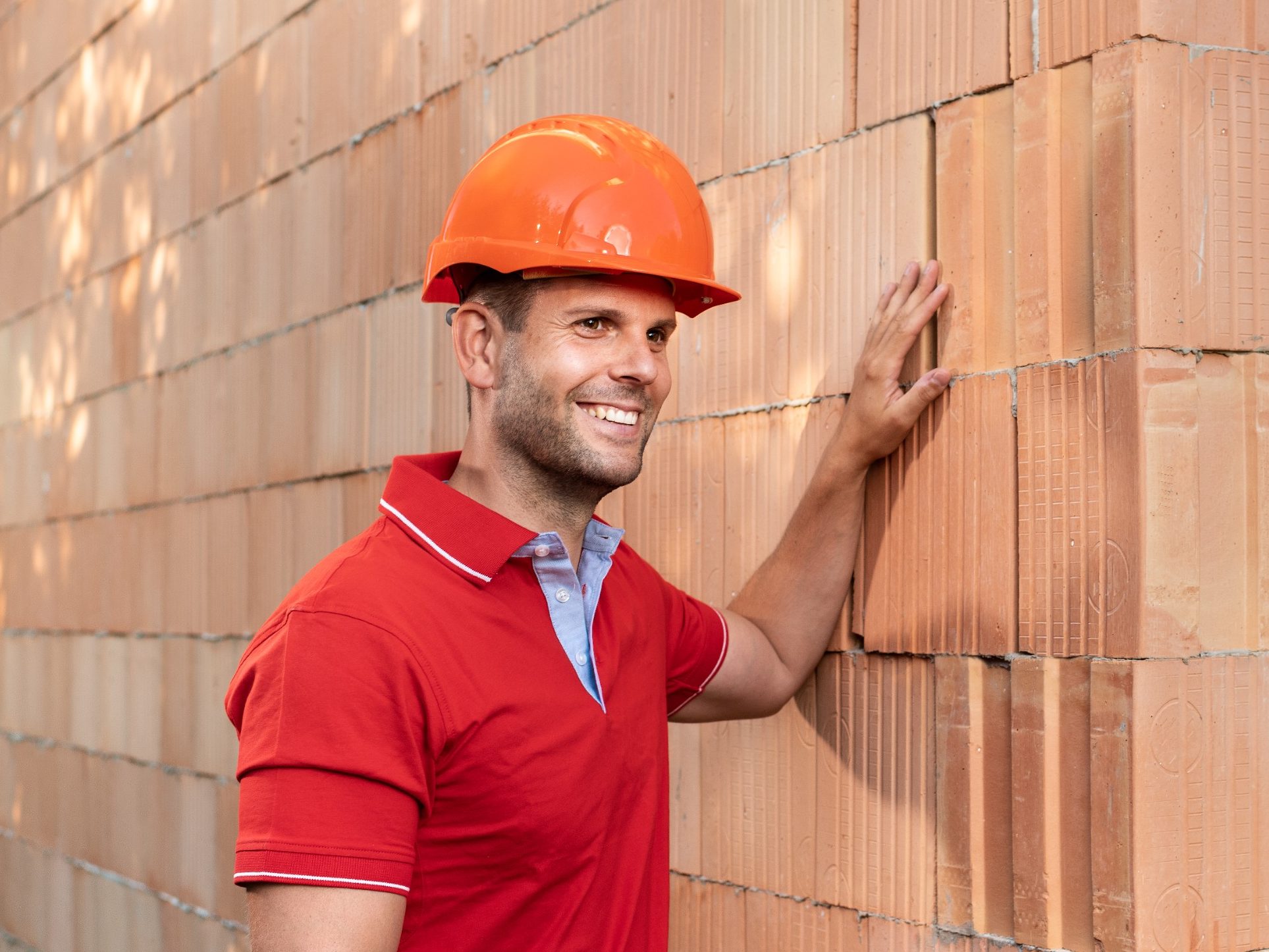 Muž stojící před rohem cihlové stěny z cihel Porotherm T Profi na hlavě s oranžovou helmou rukou hladící cihly